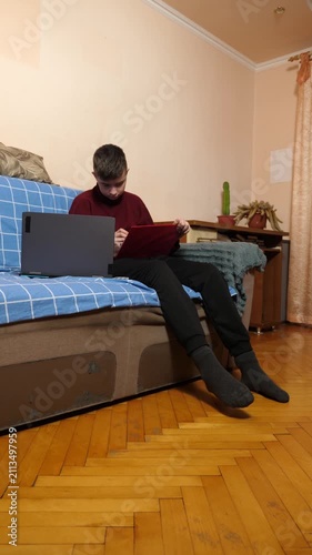 Teenager is sitting on a couch, using a tablet beside a laptop while studying or doing homework at home.