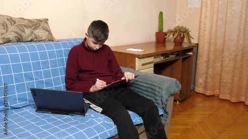 Teenager is sitting on a couch, continuously writing on a tablet with a stylus beside a laptop while studying and doing homework.