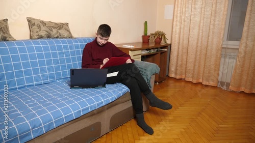 Teenager is sitting on a couch, carefully copying and sketching material from a laptop onto a tablet while studying at home.