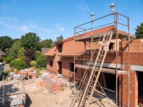 Modern two-story red brick house under construction with scaffolding, yellow ladder, and temporary fencing on sunny day.