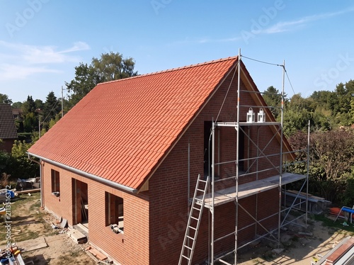 Modern two-story red brick house under construction with scaffolding, yellow ladder, and temporary fencing on sunny day.
