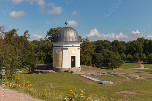 HELSINKI, FINLAND - AUGUST 14, 2025: A small yellow astronomical observatory building with a dark dome stands in a park surrounded by lawn and wildflowers.