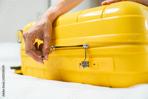 A woman's hands are zipping up a yellow wheeled suitcase after she's finished packing for her trip.