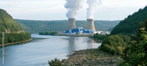 Industrial plant with cooling towers and steam plumes beside a river and forested hills
