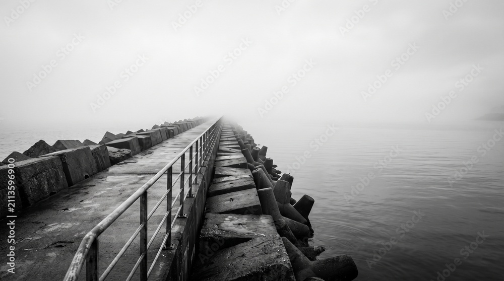 Obraz premium Concrete pier with tetrapod breakwater vanishing into thick fog at sea