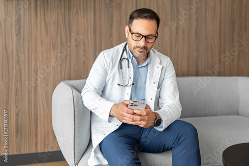 Doctor sitting on a couch, holding a smartphone, representing modern healthcare, digital communication, and telehealth services in a clinic or office setting