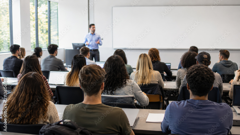 Obraz premium Students attentively listen to a male instructor presenting in a bright classroom with large windows and a whiteboard.