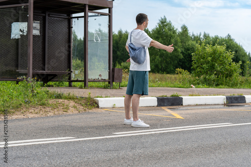 Man with a bag hitchhikes on a rural road by extending his thumb. The wide road stretches into the distance with a vehicle approaching, highlighting travel and adventure themes.