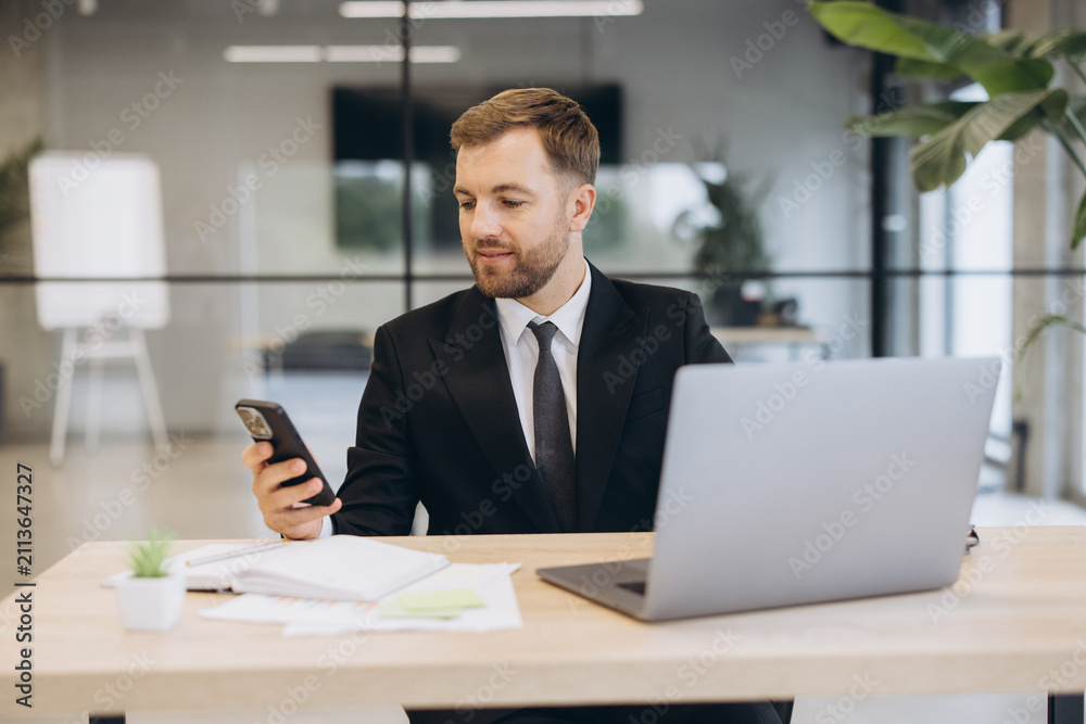 Obraz premium Businessman managing communication on smartphone while working on laptop in modern office environment