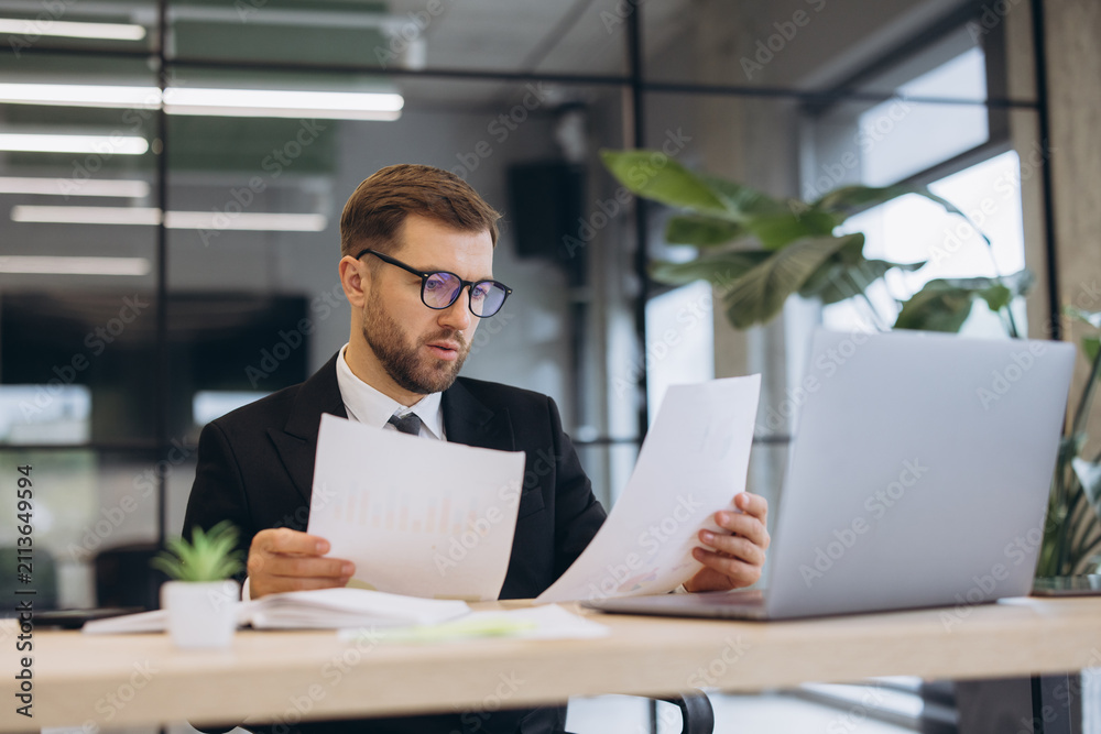 Obraz premium Young businessman reviewing financial reports in office, analyzing documents and working on laptop