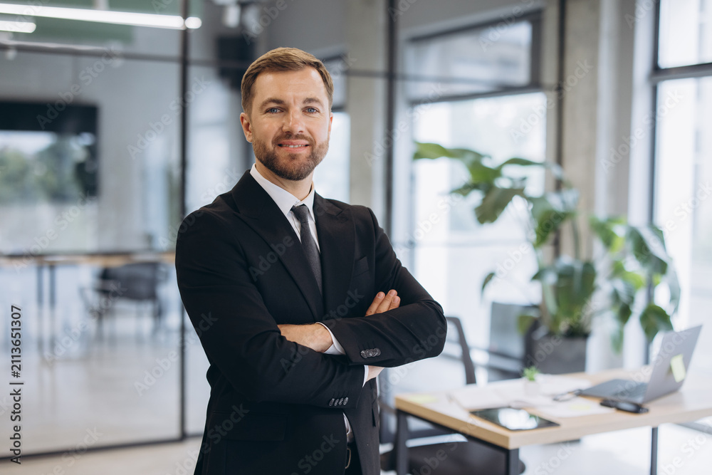 Fototapeta premium Male professional in a business suit with hands crossed, smiling at camera in a bright corporate office setting