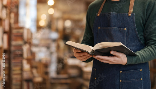 Bookseller reading and holding an open book in a warm bookstore interior. The composition has shallow depth of field and space for text on the left.