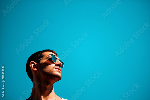 A young man stands outdoors under a bright blue sky, wearing sunglasses and enjoying the sunshine, looking upward with a relaxed expression. Summer Holidays and leisure concept. Generative AI.