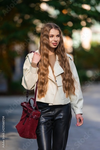 Young woman with long wavy brown hair wearing beige trench coat stands outdoors along tree lined street in daylight, carrying burgundy handbag over shoulder and smiling softly, casual fashion