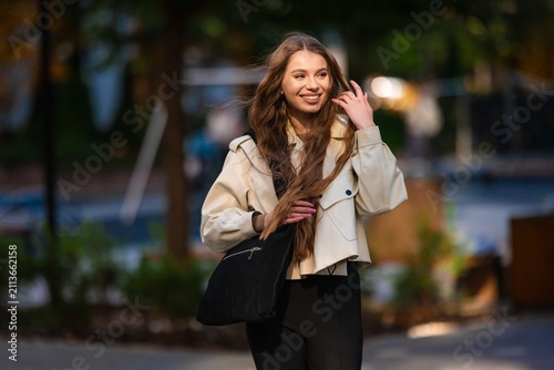 Smiling woman wearing beige trench coat outdoors park sunlight portrait warm cheerful expression natural styling fashion accessory shoulder bag casual urban setting