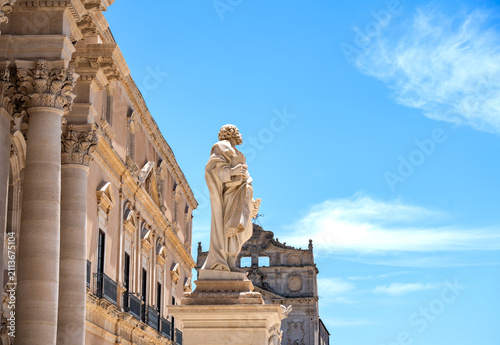 Set against a clear blue sky, the statue of Saint Peter stands majestically in front of the façade of Syracuse Cathedral