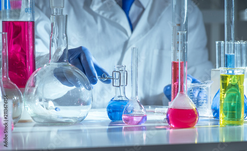 Chemistry laboratory scene with colorful chemical solutions in glass flasks and cylinders, scientist in gloves writing results, analytical experiment workspace.