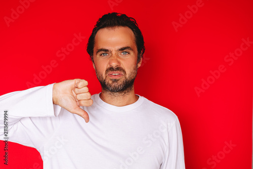 Disappointed middle aged Caucasian man showing thumbs down gesture with disgusted annoyed face expression looking at camera on bright red studio background, negative review rejection