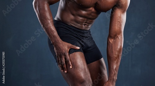 Male athlete with muscular physique poses in black shorts against a dark background, showcasing strength and fitness in a studio setting