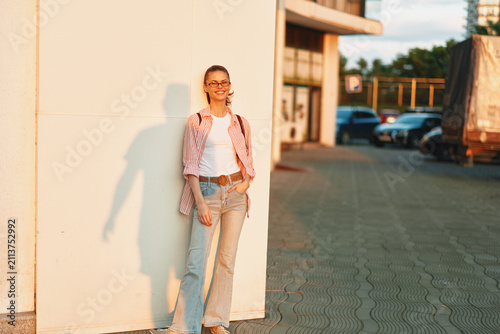 Принти на полотні A confident woman stands outdoors beside a light wall, wearing jeans and a white top with a pink plaid shirt, smiling softly in warm daylight