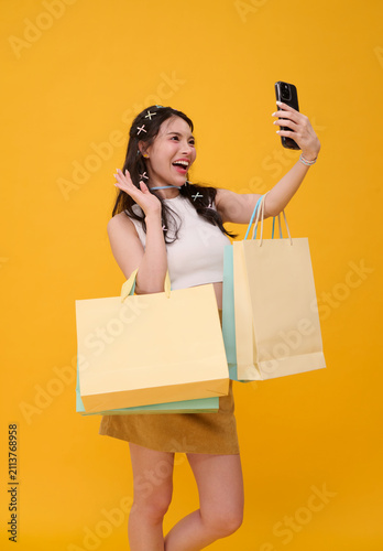 Shopping selfie with joyful Asian woman holding colourful bags and smartphone in studio expressing retail excitement social sharing consumer confidence and modern mobile lifestyle.