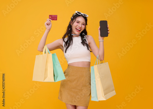 Mobile shopping payment with joyful Asian woman holding credit card smartphone and shopping bags in studio promoting cashless purchases retail convenience and digital consumer confidence.