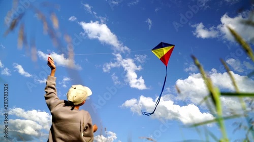 Child flies kite in open field on sunny day. Clouds drift across bright blue sky behind them. Kite dances high, colorful, joyful, free. Perfect for scenes of childhood joy, outdoor fun