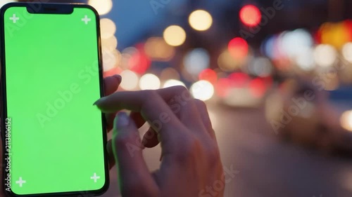 Close-up of a persons hand using a smartphone with a green screen at night, with blurred city lights in the background.
