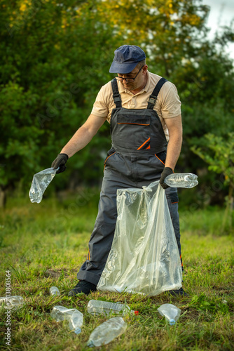 A worker collects plastic bottles and rubbish in a green field. He wears gloves and works to clean the area. The sun shines through trees in the background