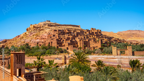 People explore Ait Benhaddou, a historic ksar in Morocco. The village shows traditional earthen clay architecture. It is set against desert hills under a blue sky.