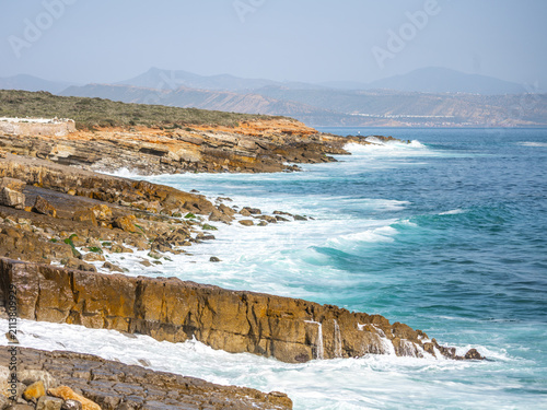 Waves crash against the rocky coast of the Atlantic Ocean in southern Morocco. The landscape shows cliffs and distant mountains under a bright sky.