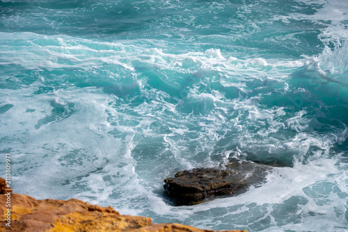 Waves roll heavily against rocks at the shore. The ocean displays high energy as water splashes and foams. The scene captures the power of nature during a stormy day.