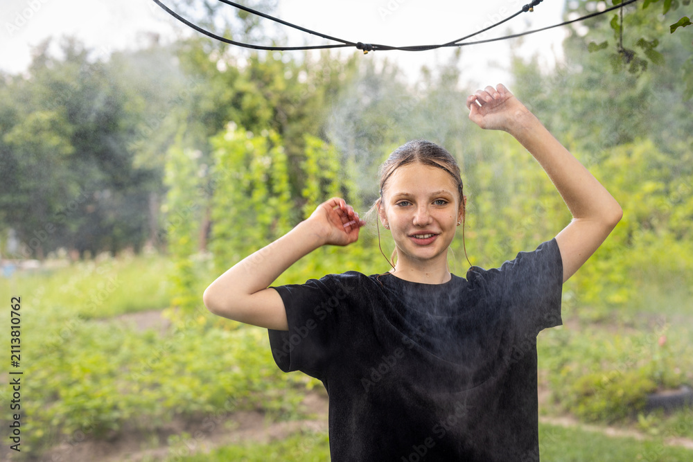 Obraz premium Teenage Girl Enjoying Cooling Water Mist in a Garden. Teenage girl in a black shirt raises her arms beneath a cooling water mist in a lush green garden.