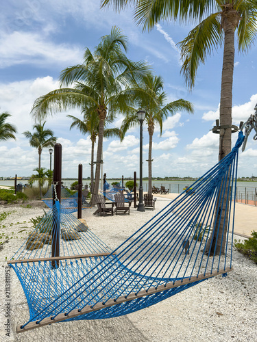 Blue mesh hammock hanging near palm trees and classic outdoor lounge chairs at a scenic point in a lakeside town center in southwest Florida. (Foreground focus.) Lifestyle and community motifs.