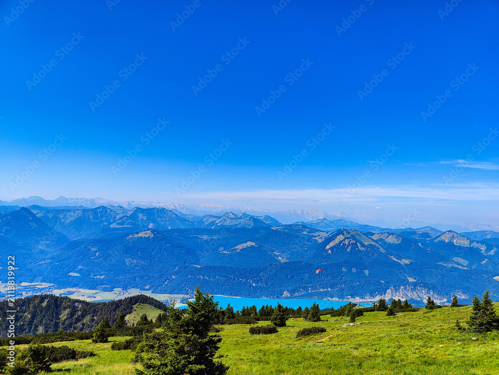 Obraz premium A vast mountain landscape unfolds from a grassy green peak, showing a bright blue lake in the valley below and a distant paraglider.