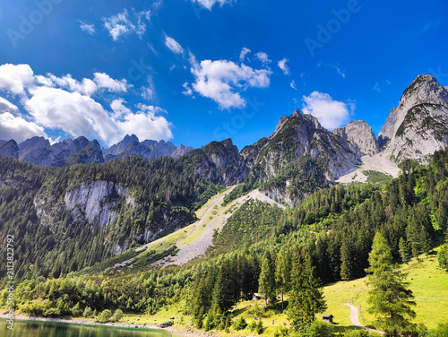 A sunny landscape showing towering mountain peaks next to a forested lake.