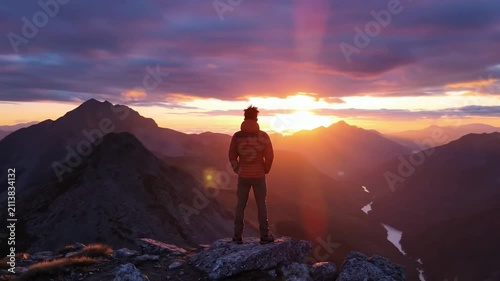Cinematic wide shot of a lone hiker standing still on a dramatic mountain peak, gazing at a vibrant purple and orange sunset to evoke themes of success, exploration, freedom, and majestic nature.