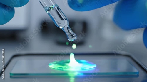 Extreme slow motion of a gloved hand using a glass pipette to drop clear liquid onto a microscope slide: a glowing chemical reaction unfolds within a sterile, professional laboratory environment.