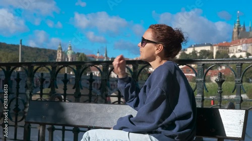 Tourist enjoying prague cityscape from a bench. Young woman tourist wearing sunglasses and casual clothes sits on a bench, enjoying the cityscape of prague with prague castle in the background