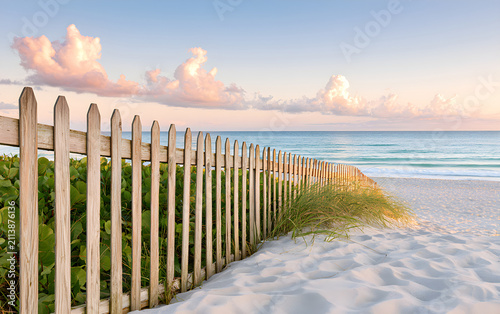Sandy beach with wooden fence and green plants under blue sky with clouds. Ocean waves gently wash shore at sunrise or sunset. Tropical coast and calm sea