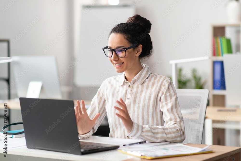 Fototapeta premium Smiling Young Mixed Race Woman Engaging In Video Call While Working At Home Office. Sitting At Desk With Laptop, Enjoying Professional Interaction In Modern Workspace. Business Concept.