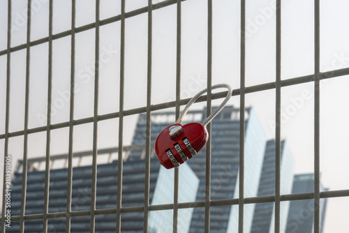 A single bright, red, heart-shaped lock with a combination is affixed to a fence on a bridge. Multiple tall skyscrapers are in the background.The love lock symbolizes commitment for two people in love