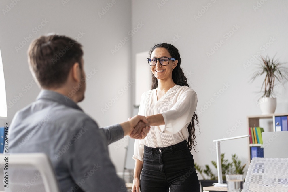 Obraz premium Smiling Hispanic Woman Engaging In Handshake With Male Colleague At Desk In Modern Office, Demonstrating Positive Interaction And Professionalism, Showing Collaboration In Business Concept.