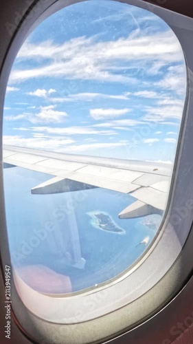 Vertical screen. Airplane wing visible from window showing ocean, islands, and clouds under a bright sky during daytime flight