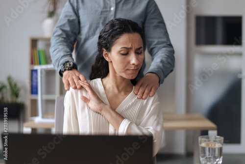 Worried Hispanic Woman In Her 30s Receiving Shoulder Massage From Male Colleague In Office. She Is Frowning  Showing Stress And Discomfort While Sitting In Front Of Laptop At Workplace.