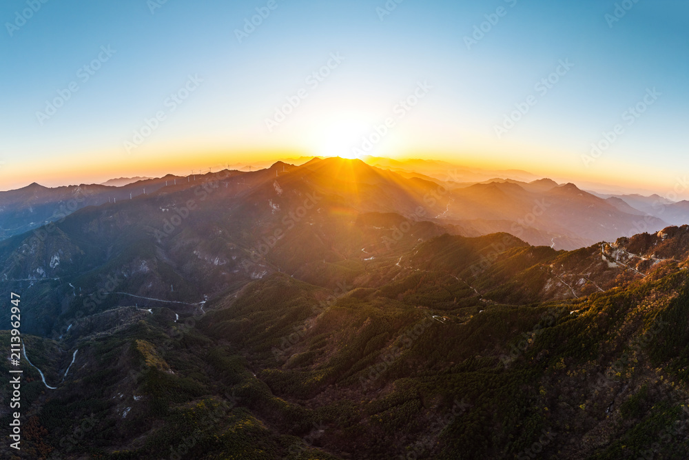 Obraz premium A panoramic view of layered mountain ranges at sunrise, with the sun peaking over the distant peaks, casting warm golden light over the rugged terrain.