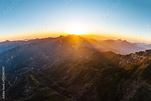 Konstfotografi A panoramic view of layered mountain ranges at sunrise, with the sun peaking over the distant peaks, casting warm golden light over the rugged terrain