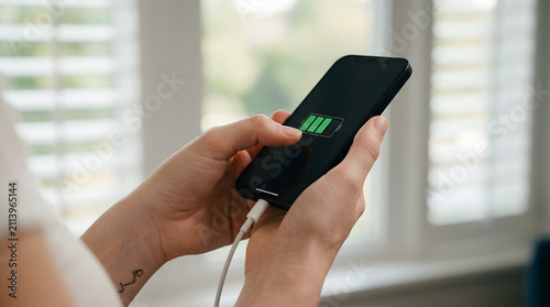 Person holding black smartphone with green charging battery icon on screen by window, mobile