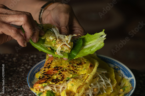 Close up of elderly hands wrapping crispy golden Vietnamese banh xeo crepe with fresh green lettuce and sprouts