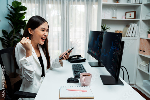Obraz na plátne Successful young Asian businesswoman earning high profit raising fist up, holding smartphone in exchange stock market's financial technology with dynamic business graph at modern office